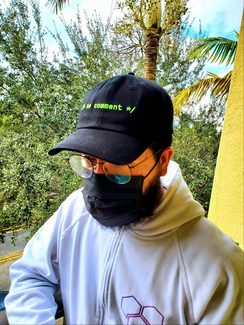 May include: A black baseball cap with a green embroidered design that says "// no comment *//". The person wearing the cap is also wearing a white hoodie with a purple and white geometric design.