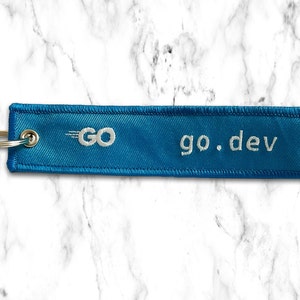 K&ouml;nnte beinhalten: Ein blauer Stoffschl&uuml;sselanh&auml;nger mit wei&szlig;en N&auml;hten und dem Text "go.dev" sowie einem wei&szlig;en Logo mit dem Text "GO" und einem wei&szlig;en stilisierten Pfeil.