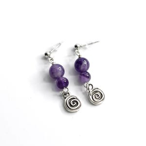 May include: A pair of dangle earrings featuring three purple beads and a silver spiral charm. The earrings have a post back and are set against a white background. The beads are a deep shade of purple.
