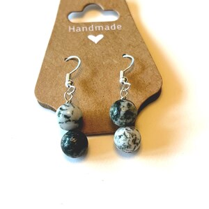 May include: Handmade earrings with two speckled stone beads in shades of black and white. The earrings hang from silver-colored hooks and are displayed on a brown card with the word "Handmade" and a heart.