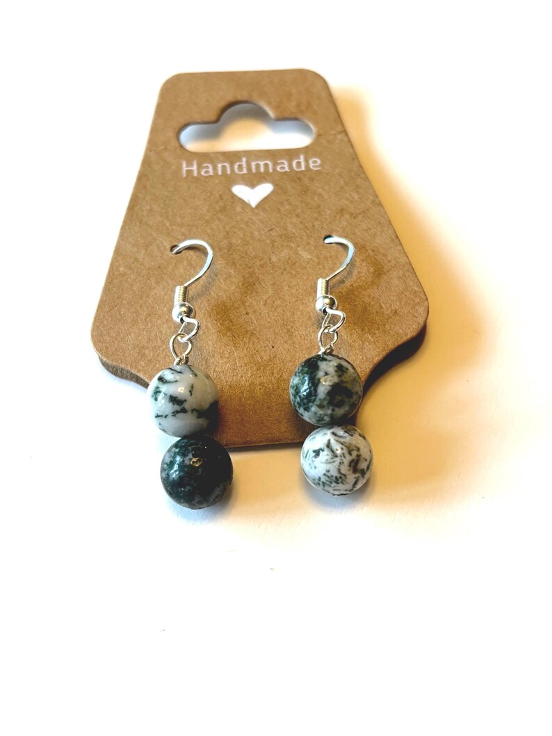 May include: Handmade earrings with two speckled stone beads in shades of black and white. The earrings hang from silver-colored hooks and are displayed on a brown card with the word "Handmade" and a heart.