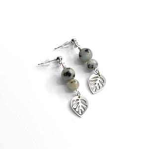 May include: A pair of dangle earrings featuring gray and white speckled beads and silver leaf charms. The earrings have a post-back closure and are set against a white background. The jewelry is a nature-inspired accessory.