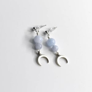 May include: A pair of dangle earrings featuring three light blue beads and a silver crescent moon charm. The earrings have a post back and are set against a white background. The earrings are a celestial-themed jewelry item.