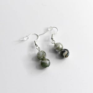 May include: A pair of dangle earrings featuring three round, marbled beads in shades of green, gray, and white. The beads are suspended from silver-colored fish hook ear wires. The earrings are displayed against a white background.