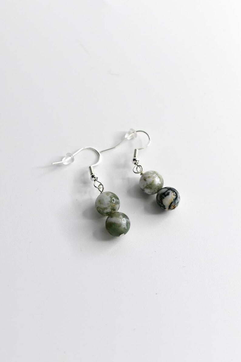 May include: A pair of dangle earrings featuring three round, marbled beads in shades of green, gray, and white. The beads are suspended from silver-colored fish hook ear wires. The earrings are displayed against a white background.