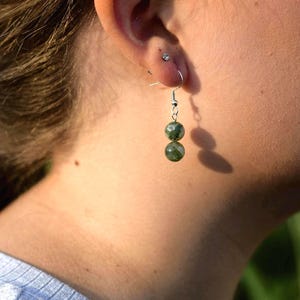 May include: A close-up of a person's ear wearing a pair of dangle earrings. The earrings feature two green, speckled beads suspended from silver-colored hooks. The person is wearing a light blue shirt.