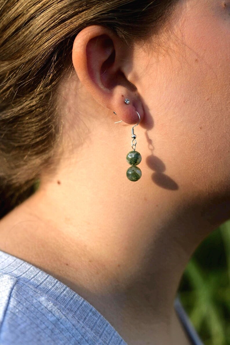 May include: A close-up of a person's ear wearing a pair of dangle earrings. The earrings feature two green, speckled beads suspended from silver-colored hooks. The person is wearing a light blue shirt.