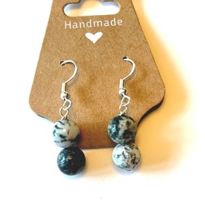 May include: Handmade earrings with silver hooks and two speckled stone beads in shades of white and dark green. The earrings hang from a brown tag with the word "Handmade" and a small heart.