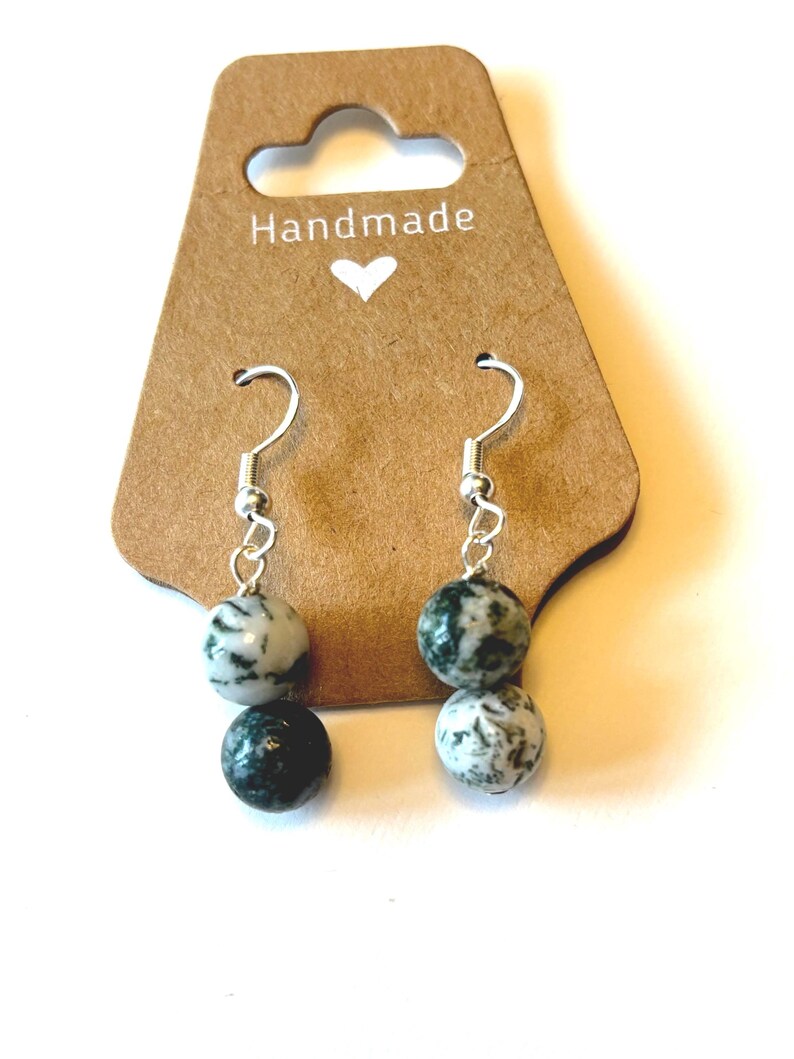 May include: Handmade earrings with silver hooks and two speckled stone beads in shades of white and dark green. The earrings hang from a brown tag with the word "Handmade" and a small heart.