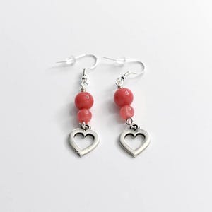 May include: A pair of dangle earrings featuring a silver heart charm and three pink beads. The earrings have a silver hook and are displayed against a white background. The earrings are a jewelry accessory.