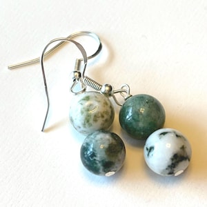 May include: A pair of dangle earrings featuring four round beads in shades of green and white. The beads are attached to silver-colored ear wires. The earrings are displayed on a white surface.