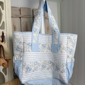 May include: A large tote bag with a light blue and white floral and striped pattern. The bag has light blue handles and accents. The bag is hanging from a white hook.