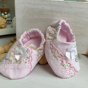 May include: A pair of pink baby booties with white bows and floral print fabric. The booties are made of soft, comfortable material.