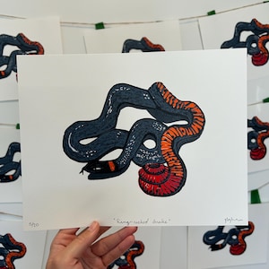 May include: A white art print featuring a dark grey snake with a bright orange ring around its neck and a vibrant red underside. The text "Ring-necked Snake" is printed at the bottom centre, with "5/20" on the left and a signature on the right.