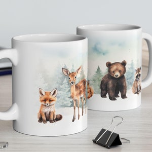May include: Two white ceramic mugs with a watercolor design of woodland animals. The mugs feature a fox, a deer, a bear, and a raccoon. The background is a light blue with white clouds and trees.
