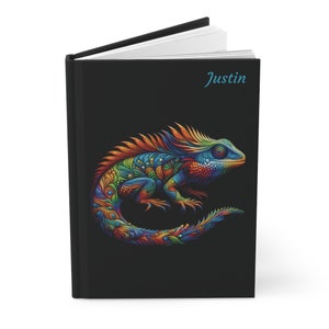May include: A black notebook with the name "Justin" printed on the front cover. The cover features a colorful, detailed illustration of a lizard with a long tail.