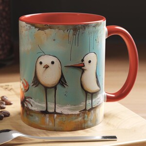 May include: A ceramic mug with a red interior and handle. The exterior features a whimsical illustration of two white birds and a crab against a teal background. The mug is sitting on a wooden surface.