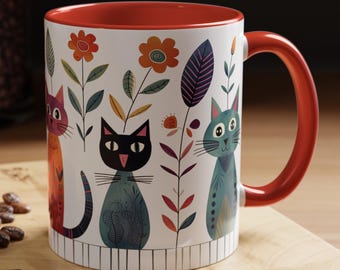 May include: A white ceramic mug with a red rim and handle. The mug features a colorful illustration of three cats with floral designs.