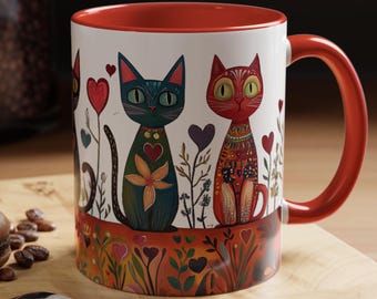 May include: A white ceramic mug with a red rim and handle. The mug features a colorful illustration of three cats with flowers and hearts. The cats are blue, red, and black.