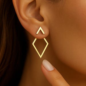 Puede incluir: Pendientes geométricos dorados. Los pendientes presentan un pequeño perno triangular y una gota en forma de diamante más grande. Las joyas se llevan en la oreja, con una vista de cerca que destaca el diseño y la forma de la oreja.