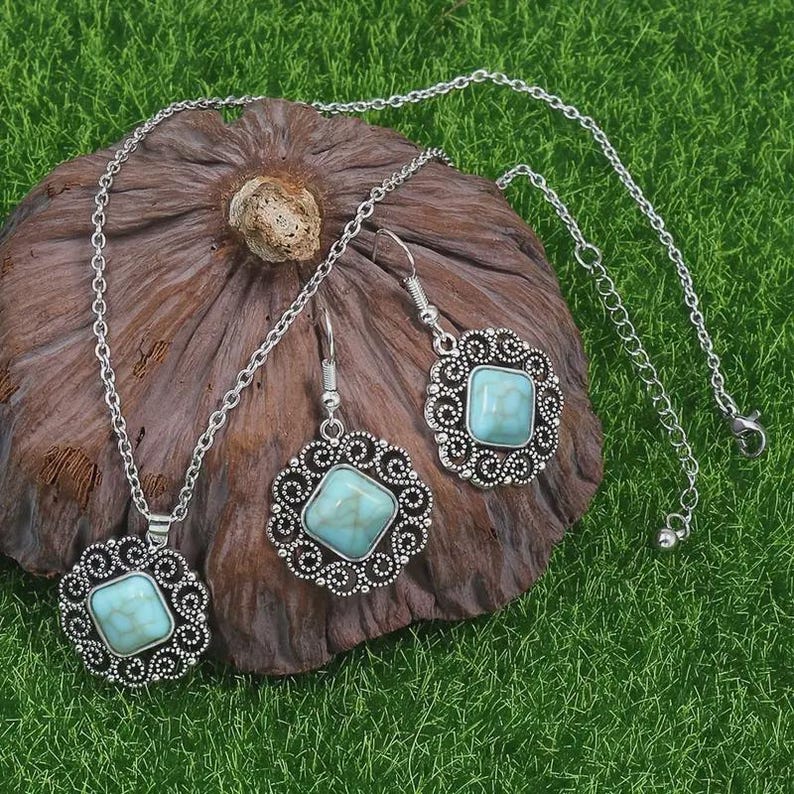K&ouml;nnte beinhalten: Ein silberfarbenes Schmuckset mit Halskette und Ohrringen. Jedes St&uuml;ck hat einen quadratischen, hellblauen Stein, der in einen dekorativen, durchbrochenen Rahmen gefasst ist. Das Set wird auf einer braunen, strukturierten Oberfl&auml;che vor gr&uuml;nem Hintergrund pr&auml;sentiert.