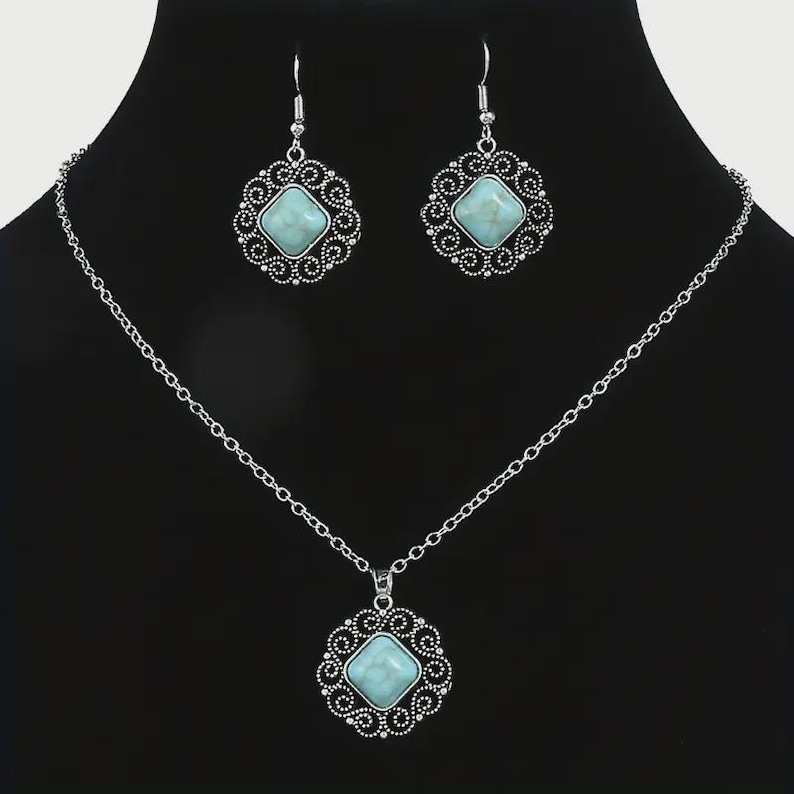 K&ouml;nnte beinhalten: Ein silberfarbenes Schmuckset mit Halskette und passenden Ohrringen. Jedes St&uuml;ck hat einen quadratischen, hellblauen Stein, der in einem dekorativen, durchbrochenen Rahmen gefasst ist. Die Halskette hat eine zarte Kette.