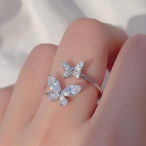 May include: A silver-toned ring featuring two butterfly designs. One butterfly is positioned on top, while the other is on the side, both adorned with clear, sparkling gemstones. The ring is designed to wrap around the finger.