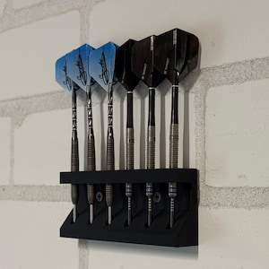 May include: A black dart holder mounted on a white brick wall, holding six darts. The darts have black and silver shafts with various flight designs, including blue and black patterns. The holder is made of black plastic.