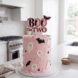 May include: A cylindrical cake with light pink frosting decorated with white ghost shapes, black bat cutouts, and small silver sprinkles. A black cake topper reads "BOO I'm TWO" with a small bat silhouette. The cake sits on a white pedestal stand.