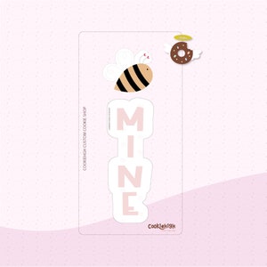 May include: A pink cookie cutter shaped like the word "MINE" with a bee and a donut on top. The bee is yellow and black with wings. The donut is brown with white frosting and sprinkles. The cookie cutter is on a white background with a pink border.