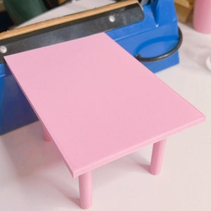 May include: A pink table with four legs. The table is made of plastic and has a smooth surface. The table is small and would be suitable for a dollhouse or other miniature setting.