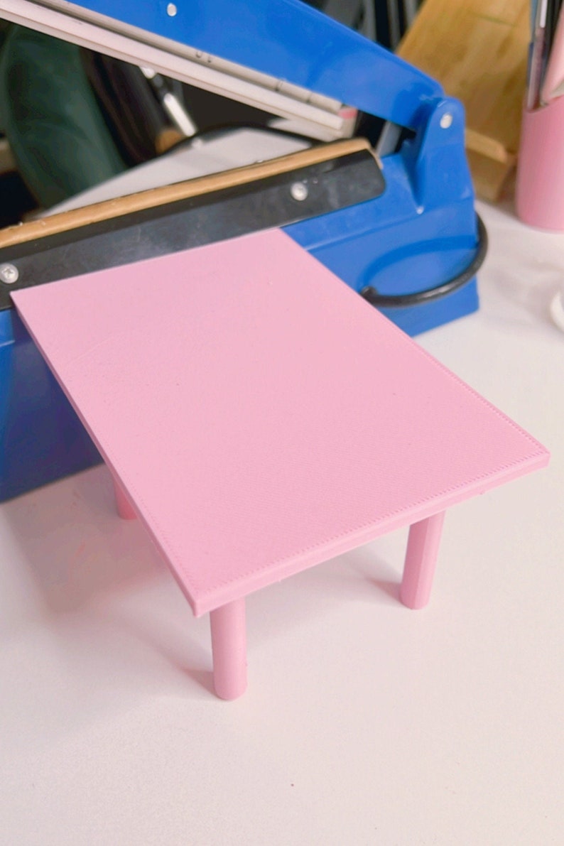 May include: A pink table with four legs. The table is made of plastic and has a smooth surface. The table is small and would be suitable for a dollhouse or other miniature setting.
