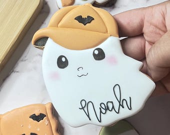 Op de afbeelding: Een verzameling Halloween-koekjes. Het belangrijkste koekje is een wit spook met een oranje hoed met een zwarte vleermuis en de naam "Noah" in zwart schrift. Andere koekjes zijn een "Hey Boo" koekje en een snoepmaïskoekje.