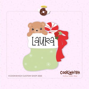 May include: A Christmas stocking-shaped cookie cutter with the name "LAURA" on a white banner. A brown bear peeks over the banner. The stocking is light green with white snowflakes, a red bow, and a candy cane. A donut with wings is at the top.
