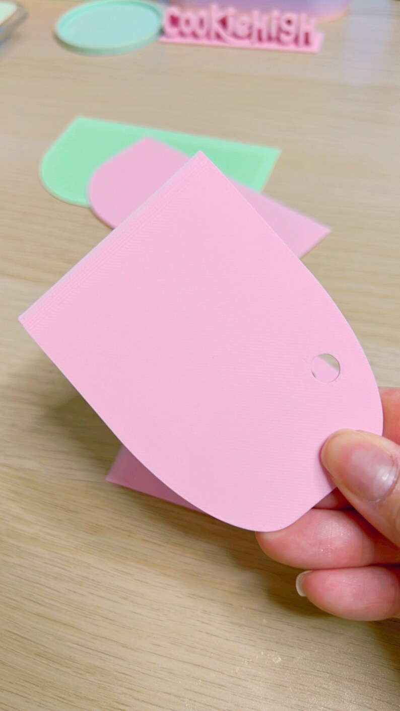 May include: A pink plastic scraper with a hole in the handle. The scraper is used for spreading frosting or icing on cakes and cookies.