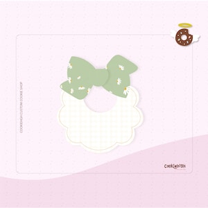 May include: A white and green cookie cutter shaped like a baby bib with a bow on top. The bow has a green background with white daisies. The cookie cutter is on a pink background with white dots.