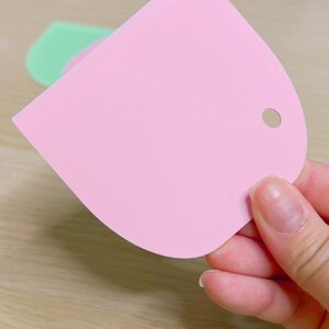 May include: A pink plastic scraper with a rounded edge and a hole in the top. The scraper is used for spreading frosting or icing on cakes.