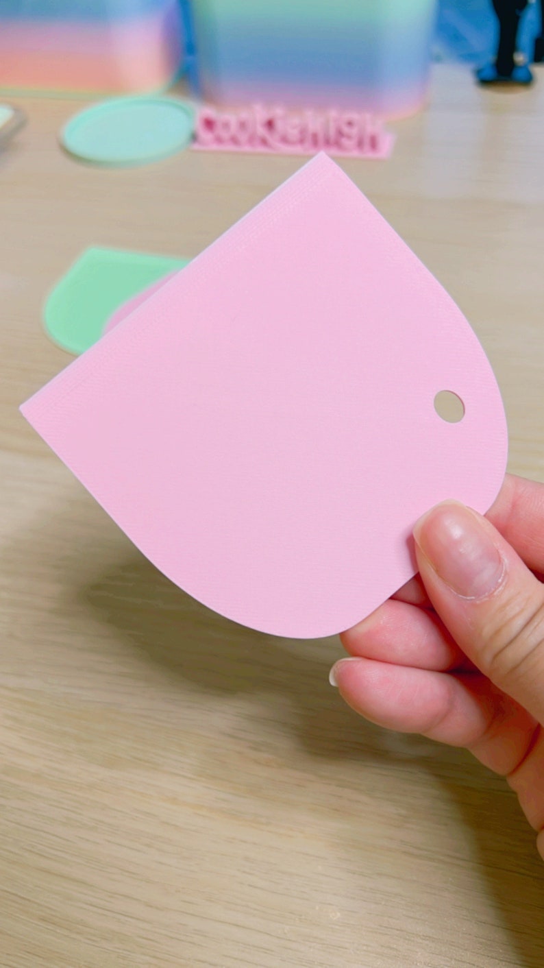 May include: A pink plastic scraper with a rounded edge and a hole in the top. The scraper is used for spreading frosting or icing on cakes.