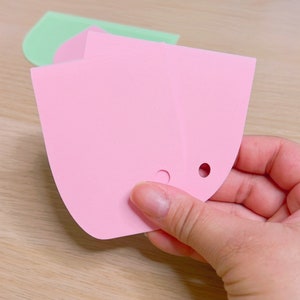 May include: Two pink silicone baking tools with rounded edges and holes. The tools are held in a hand.