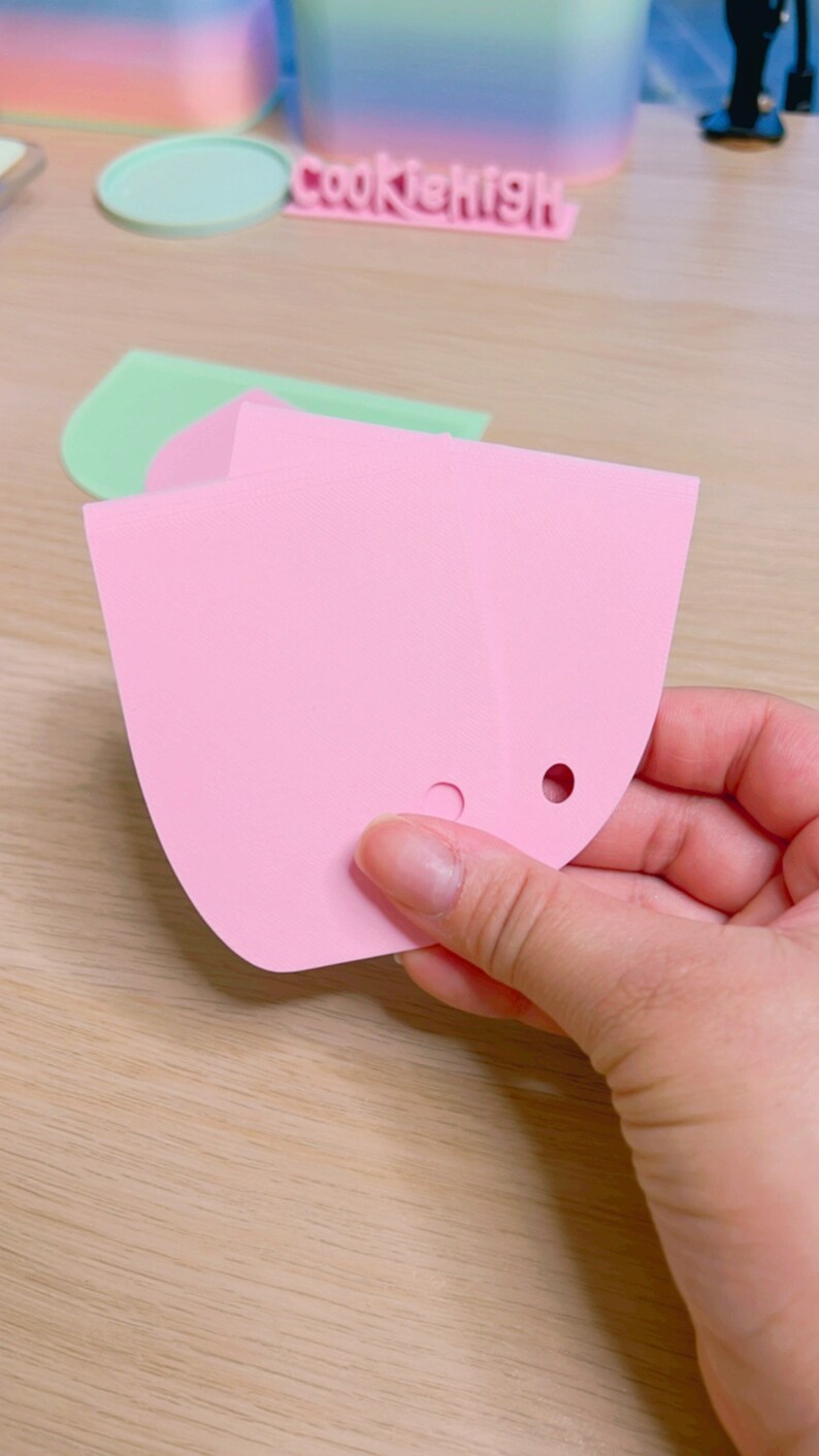 May include: Two pink silicone baking tools with rounded edges and holes. The tools are held in a hand.