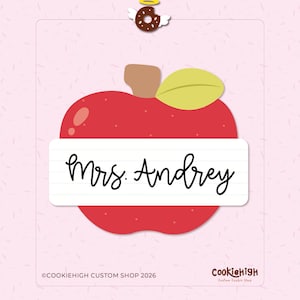 May include: A red apple-shaped cookie cutter with a white rectangle in the center, inscribed with "Mrs. Andrey" in cursive. A brown stem and a yellow-green leaf are attached. A donut with wings and a halo is at the top. The background is pink.