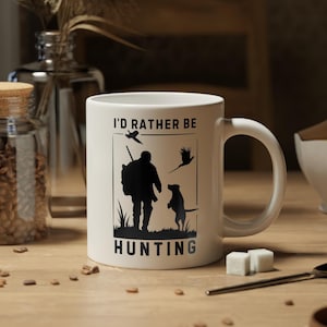 May include: White ceramic coffee mug with a black silhouette of a hunter with a dog and the text "I'd rather be hunting".