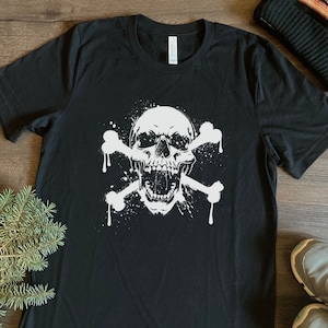 Könnte beinhalten: Schwarzes T-Shirt mit einem weißen Totenkopf- und Knochen-Motiv. Der Totenkopf hat einen offenen Mund und einen Spritz-Effekt.