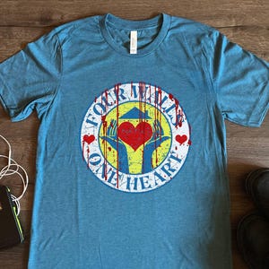 Op de afbeelding: Een blauw T-shirt met een witte cirkelvormige afbeelding waarop "Four Walls One Heart" staat in een rood-wit-geel kleurenschema. De afbeelding bevat ook een rood hart en twee handen die ernaar reiken.