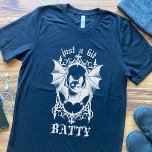 May include: Dark navy blue t-shirt with a white graphic of a bat inside an ornate frame, with the words "just a bit ratty" in a decorative font. The shirt is on a wooden surface with a beanie, phone, earbuds, and shoes.