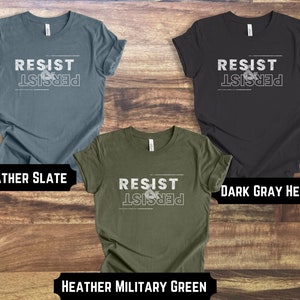 May include: Three t-shirts with the text "Resist & Persist" in white lettering on a dark background. The t-shirts are in the colors heather slate, dark gray heather, and heather military green.