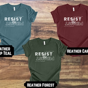 May include: Three t-shirts in different colors with the text "Resist & Persist" printed on the front. The colors are heather deep teal, heather cardinal, and heather forest.