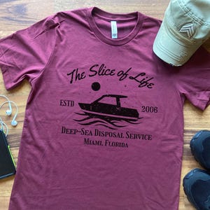 以下が含まれることがあります： 「The Slice of Life」という文字の上にボートが描かれた、マルーンのTシャツ。ボートの下には「Deep-Sea Disposal Service Miami, Florida」という文字が書かれている。また、「ESTD 2006」という文字も含まれている。