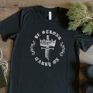 Pode incluir: Camiseta preta com um design gráfico branco de uma coroa e uma espada com o texto "Be Strong Carry On".