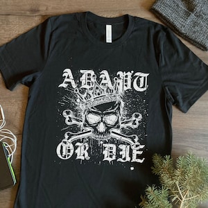 May include: Black t-shirt featuring a skull wearing a crown, with crossbones and the words "ADAPT OR DIE" in a gothic font. The design is in white, with a distressed, splattered effect. A beanie, earbuds, and a phone are in the background.